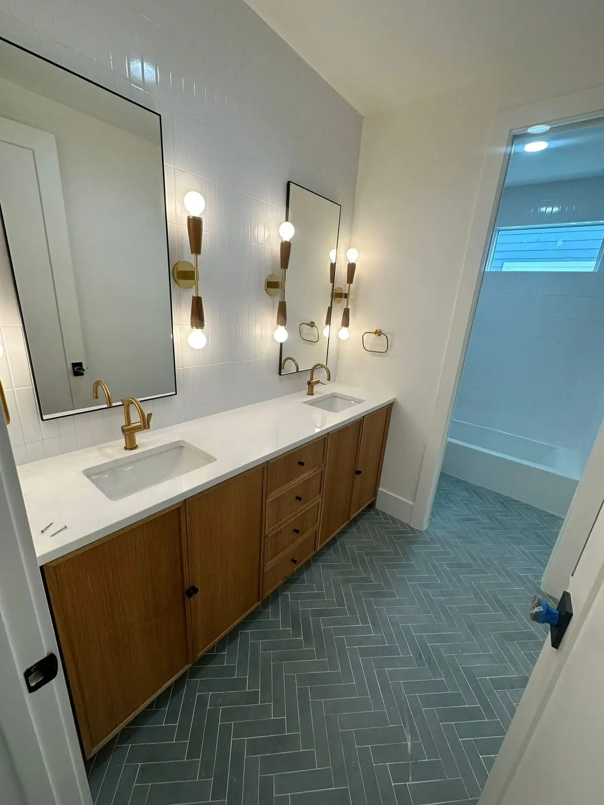 Wood double vanity with a white quartz top, brass taps and black-framed mirrors, above a teal herringbone tile floor