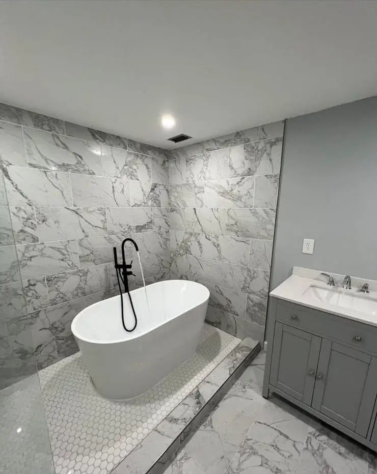 Bathroom in marble-look porcelain with a freestanding tub standing on a hexagonal mosaic platform, black tapware and a grey vanity alongside