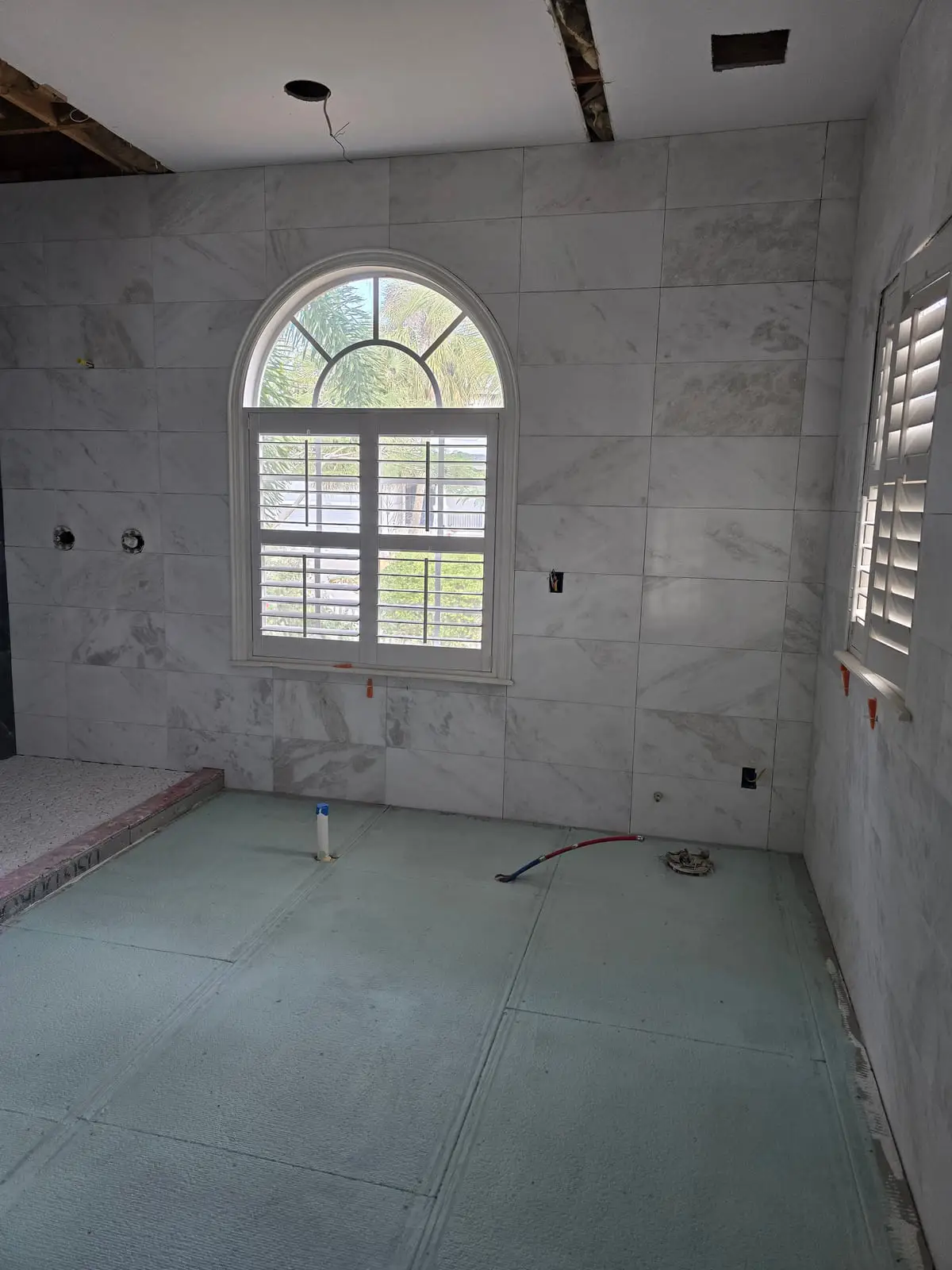 Bathroom mid-installation with white marble-look walls set around an arched window and the floor still on bare backer board
