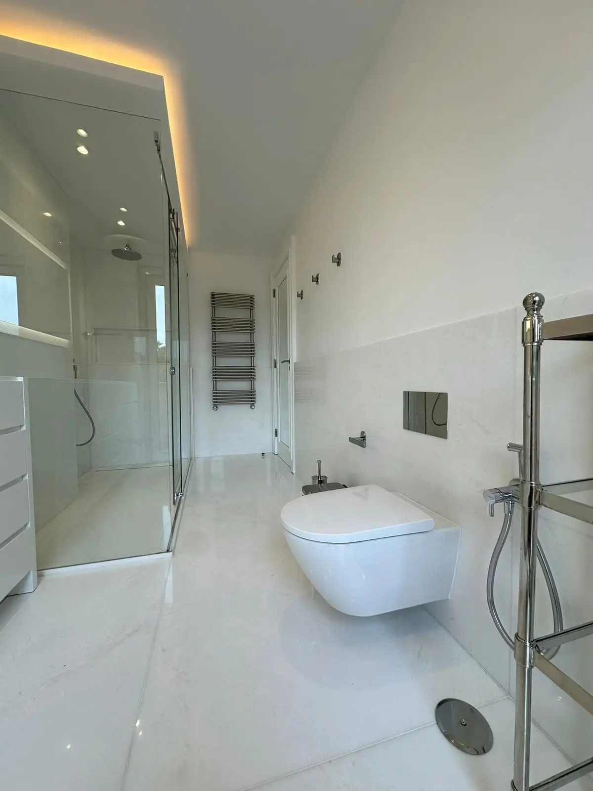 Bathroom finished in white marble-look porcelain with a glass shower enclosure, wall-hung toilet, chrome towel radiator and a cove-lit ceiling